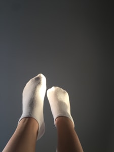 Two feet wearing white socks are raised against a plain, dark background. The socks and the feet are lit by a light source, creating a contrast with the muted background.