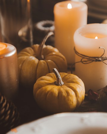 A cozy autumnal scene features several mini pumpkins surrounded by lit candles. The candles vary in size and are warmly glowing, creating a gentle and inviting atmosphere. One candle is wrapped with a rustic twine, adding a touch of rustic charm.