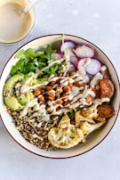 A vibrant bowl filled with a variety of healthy ingredients such as quinoa, roasted chickpeas, avocado slices, arugula, radish slices, roasted cauliflower, and sweet potatoes. The dish is drizzled with a creamy dressing. A separate bowl with additional dressing and a spoon is placed nearby.
