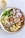 A vibrant bowl filled with a variety of healthy ingredients such as quinoa, roasted chickpeas, avocado slices, arugula, radish slices, roasted cauliflower, and sweet potatoes. The dish is drizzled with a creamy dressing. A separate bowl with additional dressing and a spoon is placed nearby.