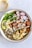 A vibrant bowl filled with a variety of healthy ingredients such as quinoa, roasted chickpeas, avocado slices, arugula, radish slices, roasted cauliflower, and sweet potatoes. The dish is drizzled with a creamy dressing. A separate bowl with additional dressing and a spoon is placed nearby.