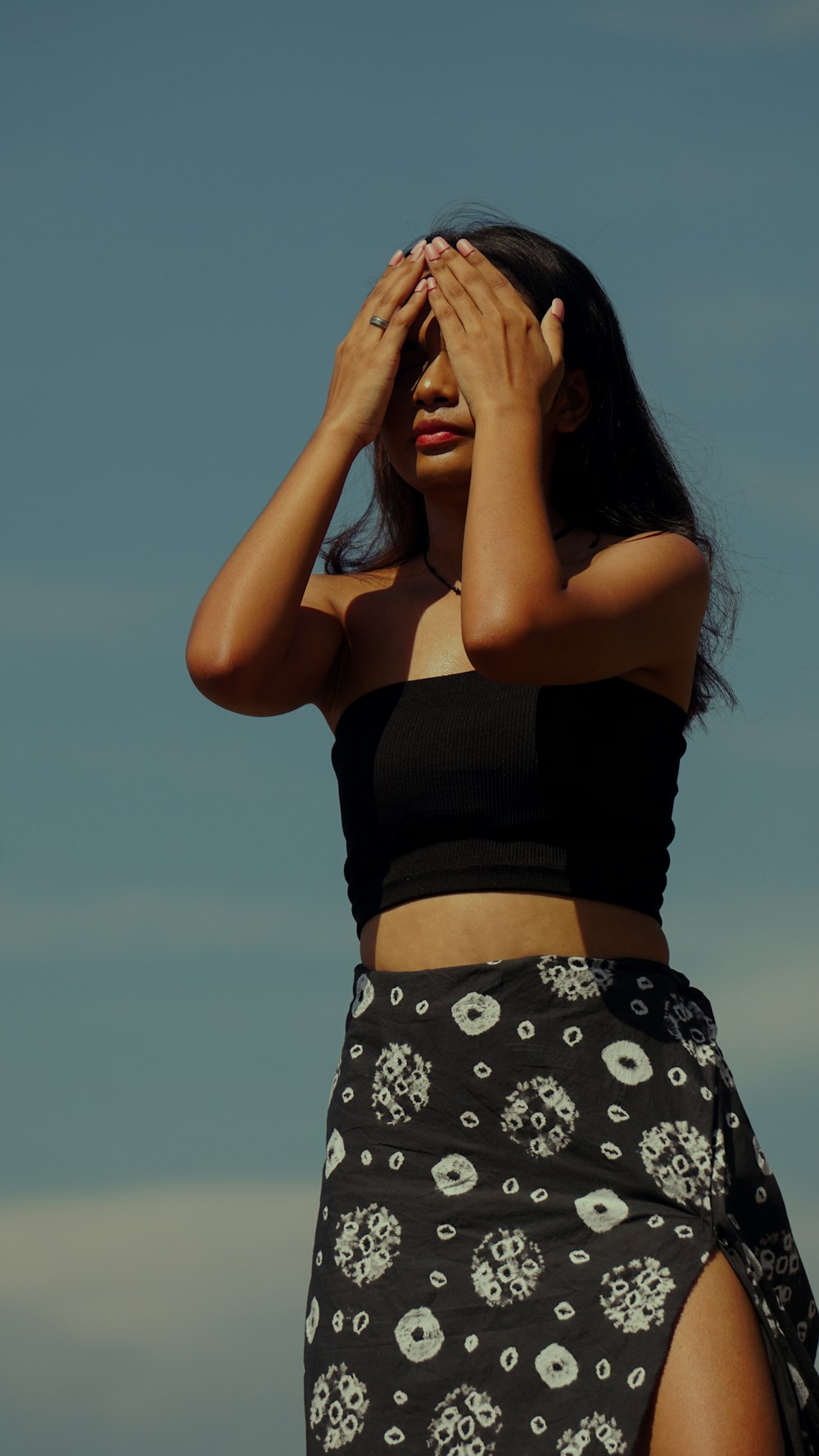 woman in black tank top and white floral skirt covering her face with her hand