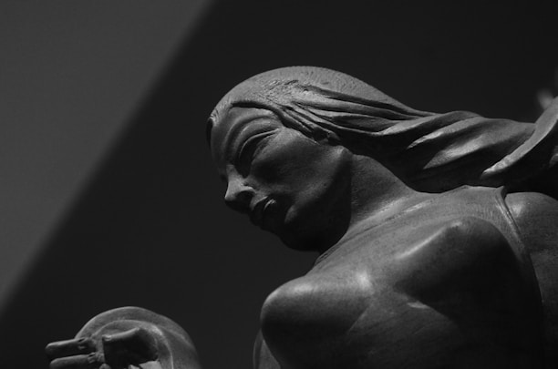 A close-up shot of a detailed sculpture depicting a person with long flowing hair, captured in a monochromatic tone. The sculpture appears to be made of a dark material, emphasizing the contours and expressions on the figure's face and body.