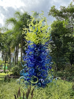 A vibrant glass sculpture with twisting, tentacle-like forms in shades of blue and green stands amongst lush greenery. The translucent structure appears organic yet intricate, resembling sea life or a fantastical plant, and is set in a garden with palm trees under a cloudy sky.