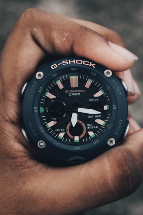 A black G-Shock Casio wristwatch is being held in a hand, showcasing its detailed analog and digital display. The watch face features multiple smaller dials and hands, with markings highlighted in a combination of green, pink, and metallic accents. The robust design and prominent branding emphasize its durability and style.