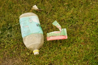 An empty plastic bottle and a piece of torn packaging are lying on the grass. The bottle is large with a faded, light blue label, and the packaging appears to be multi-colored, primarily green and red. The surrounding area is covered with grass and small plants.