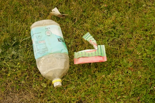 An empty plastic bottle and a piece of torn packaging are lying on the grass. The bottle is large with a faded, light blue label, and the packaging appears to be multi-colored, primarily green and red. The surrounding area is covered with grass and small plants.