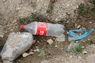 A discarded plastic bottle is lying on rocky and earthy ground, next to a blue plastic piece and surrounded by gravel and small patches of grass. A large stone is nearby, partially covered with dirt.