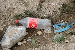 A discarded plastic bottle is lying on rocky and earthy ground, next to a blue plastic piece and surrounded by gravel and small patches of grass. A large stone is nearby, partially covered with dirt.