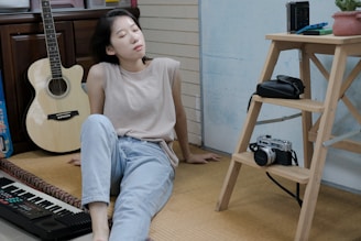A person is sitting on the floor in a relaxed pose, with eyes closed and head tilted back. They are wearing a beige sleeveless top and light blue jeans. To the left, there is an acoustic guitar resting against a wooden cabinet and an electronic keyboard nearby. On the right, a wooden step ladder holds a vintage camera, a black bag, and a small potted plant. The setting appears to be a casual indoor environment with a mix of creative and personal items.