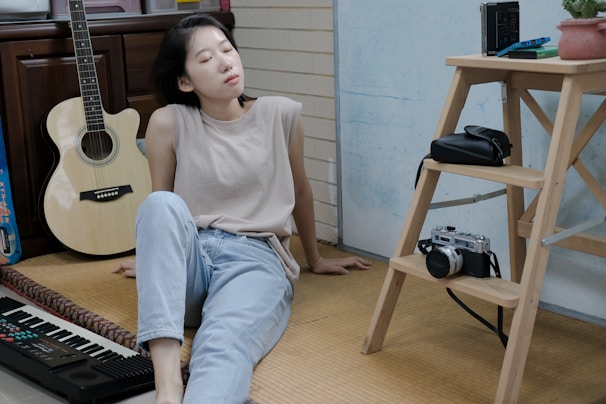 A person is sitting on the floor in a relaxed pose, with eyes closed and head tilted back. They are wearing a beige sleeveless top and light blue jeans. To the left, there is an acoustic guitar resting against a wooden cabinet and an electronic keyboard nearby. On the right, a wooden step ladder holds a vintage camera, a black bag, and a small potted plant. The setting appears to be a casual indoor environment with a mix of creative and personal items.