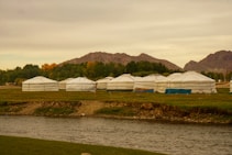 Several traditional yurts, circular and white, are set on a grassy landscape surrounded by mountains and trees. A calm river flows in the foreground, and the sky is overcast with a hint of evening light, casting a warm glow over the scene. The atmosphere is serene and remote, hinting at a nomadic lifestyle.