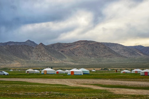 Several traditional yurts are scattered across a grassy plain with a backdrop of rugged mountains. The sky is overcast, creating a moody atmosphere. A lone car is parked on the left side, indicating a blend of traditional and modern elements.