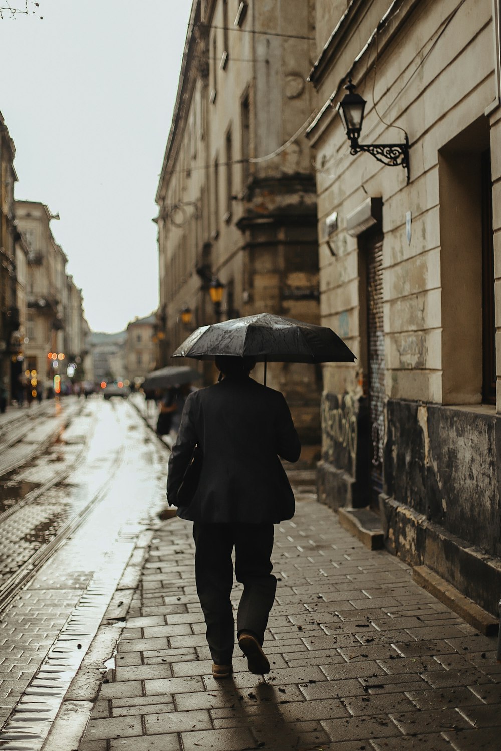 Person im schwarzen Mantel mit Regenschirm, die tagsüber auf dem Bürgersteig geht