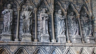 A series of stone statues are set in niches along a medieval-style wall, each figure carved in detailed drapery and individual poses. The backdrop consists of stone arches and a textured, weathered stone surface that evokes a historical or religious setting.