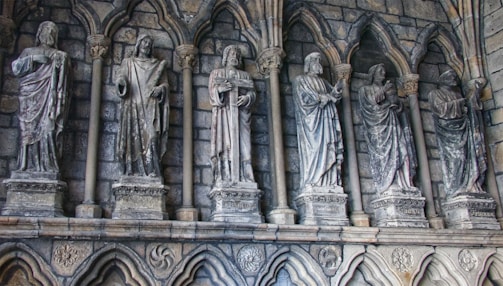 A series of stone statues are set in niches along a medieval-style wall, each figure carved in detailed drapery and individual poses. The backdrop consists of stone arches and a textured, weathered stone surface that evokes a historical or religious setting.