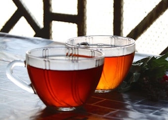 Two glass cups filled with amber-colored tea are placed on a table, accompanied by green leaves and a small red flower. The background shows a pattern of latticework.