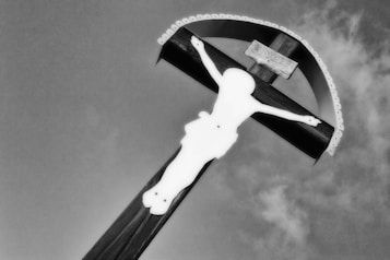 A monochromatic image featuring a wooden cross with a stylized silhouette of a figure placed centrally against the sky. The cross is topped with a decorative arch, and a small plaque is visible near the top. The sky appears cloudy and gray, adding to the dramatic impact of the scene.