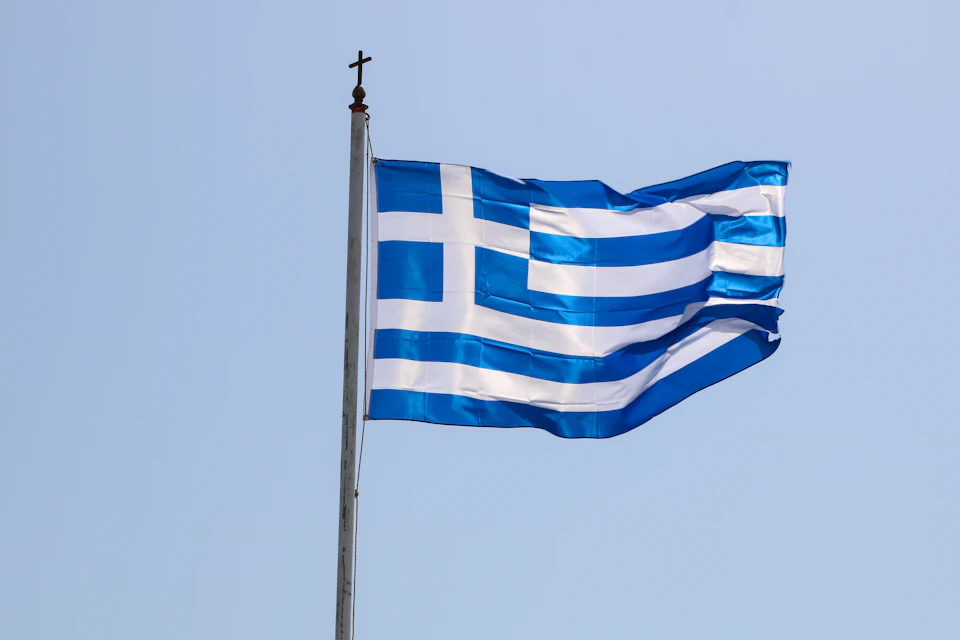 Greek flag flying high against clear blue sky with pole on left side displayed prominently