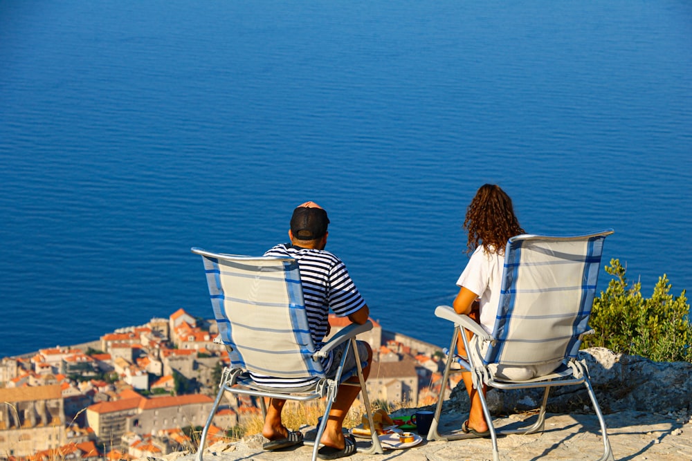 2 women sitting on white folding chairs near body of water during daytime