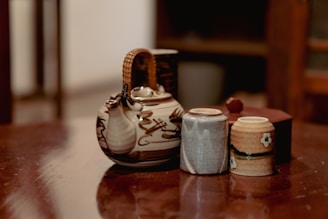 A rustic ceramic teapot with a woven handle is positioned on a wooden surface alongside two small cups. The teapot features a natural, earthy design, while the cups have subtle patterns and a flower motif. The background is slightly out of focus, enhancing the warm and cozy ambiance.