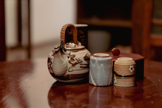 A rustic ceramic teapot with a woven handle is positioned on a wooden surface alongside two small cups. The teapot features a natural, earthy design, while the cups have subtle patterns and a flower motif. The background is slightly out of focus, enhancing the warm and cozy ambiance.