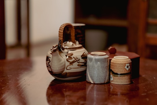 A rustic ceramic teapot with a woven handle is positioned on a wooden surface alongside two small cups. The teapot features a natural, earthy design, while the cups have subtle patterns and a flower motif. The background is slightly out of focus, enhancing the warm and cozy ambiance.
