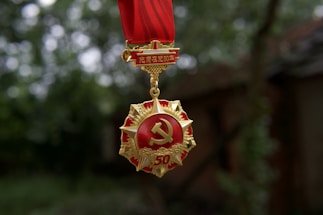A gold and red emblem with a hammer and sickle symbol hangs from a red ribbon. The medal has a star shape with intricate detailing and the number 50. The background is a blurry outdoor setting with green foliage.