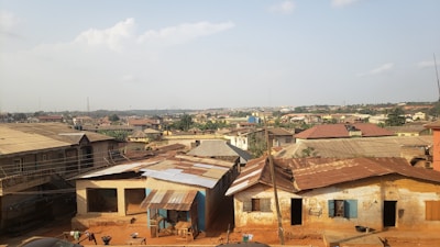A sprawling residential area filled with small, modest houses with rusty metal roofs and faded exteriors, spread across an expansive landscape. The foreground features more close-up details of the houses, including worn walls and makeshift extensions. The houses appear weathered and the dirt paths indicate a lack of pavement.