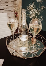 A tabletop arrangement featuring a bottle of Perrier-Jouët champagne and two filled champagne flutes placed on a geometric mirrored tray. Delicate flowers in the background add an elegant touch to the setting.