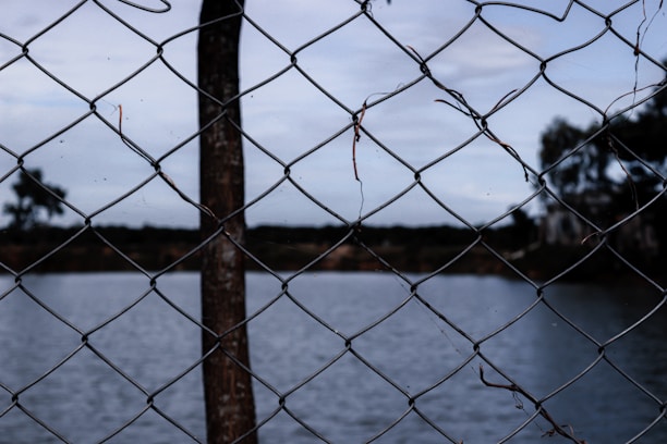 A close-up view of a chain-link fence with a blurry background that includes a body of water and trees. The fence is made of metal wire with a rustic, weathered appearance.