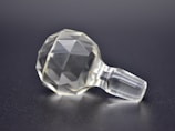 A transparent, faceted object resembling a finely cut gemstone or crystal, resting on a smooth, dark surface. The object has a geometric, spherical form with numerous facets that reflect light, giving a sense of clarity and precision.