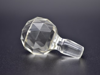 A transparent, faceted object resembling a finely cut gemstone or crystal, resting on a smooth, dark surface. The object has a geometric, spherical form with numerous facets that reflect light, giving a sense of clarity and precision.