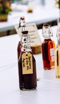 Several glass bottles with flip-top lids, filled with dark and amber liquids, are arranged on a table. The bottles have handwritten labels, one of which reads 'Blackberry & Sloe Gin Aug 2021'. The setting appears to be a bright, possibly outdoor or market-like environment with blurred elements in the background.