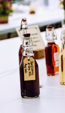 Several glass bottles with flip-top lids, filled with dark and amber liquids, are arranged on a table. The bottles have handwritten labels, one of which reads 'Blackberry & Sloe Gin Aug 2021'. The setting appears to be a bright, possibly outdoor or market-like environment with blurred elements in the background.