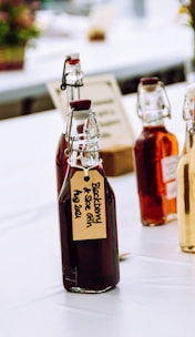 Several glass bottles with flip-top lids, filled with dark and amber liquids, are arranged on a table. The bottles have handwritten labels, one of which reads 'Blackberry & Sloe Gin Aug 2021'. The setting appears to be a bright, possibly outdoor or market-like environment with blurred elements in the background.