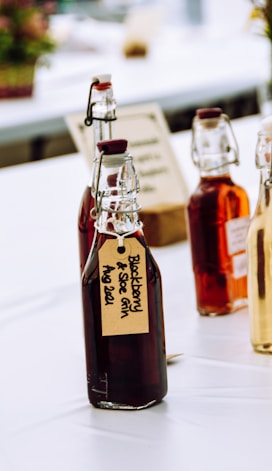 Several glass bottles with flip-top lids, filled with dark and amber liquids, are arranged on a table. The bottles have handwritten labels, one of which reads 'Blackberry & Sloe Gin Aug 2021'. The setting appears to be a bright, possibly outdoor or market-like environment with blurred elements in the background.
