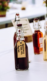Several glass bottles with flip-top lids, filled with dark and amber liquids, are arranged on a table. The bottles have handwritten labels, one of which reads 'Blackberry & Sloe Gin Aug 2021'. The setting appears to be a bright, possibly outdoor or market-like environment with blurred elements in the background.
