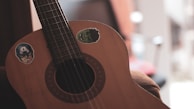 An acoustic guitar with stickers on its body, featuring cartoonish images. The background is softly blurred, suggesting an indoor setting with muted lighting.