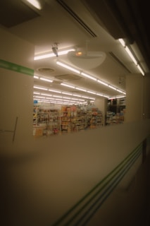 The image depicts the interior of a store, likely a convenience store or small supermarket. Shelves filled with various products are visible under a ceiling with bright fluorescent lights. The image has a warm, muted tone, suggesting it might have been taken in the evening or through tinted glass.