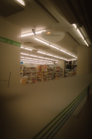 The image depicts the interior of a store, likely a convenience store or small supermarket. Shelves filled with various products are visible under a ceiling with bright fluorescent lights. The image has a warm, muted tone, suggesting it might have been taken in the evening or through tinted glass.