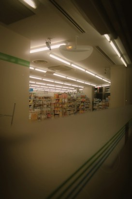 The image depicts the interior of a store, likely a convenience store or small supermarket. Shelves filled with various products are visible under a ceiling with bright fluorescent lights. The image has a warm, muted tone, suggesting it might have been taken in the evening or through tinted glass.