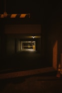 A dimly lit underground parking area with a narrow entrance. The walls are concrete, and there's a striped barrier hanging from above. Two traffic cones are visible on the right side.