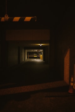 A dimly lit underground parking area with a narrow entrance. The walls are concrete, and there's a striped barrier hanging from above. Two traffic cones are visible on the right side.
