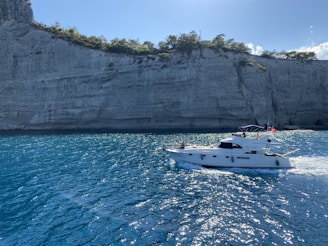 A motor yacht is cruising on a vibrant blue sea against the backdrop of a steep cliff with sparse greenery. The water is sparkling under the sunlight, adding a sense of freshness and openness.