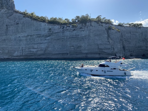 A motor yacht is cruising on a vibrant blue sea against the backdrop of a steep cliff with sparse greenery. The water is sparkling under the sunlight, adding a sense of freshness and openness.