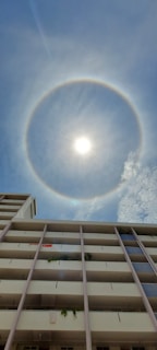 A bright sun is encircled by a distinct halo against a clear blue sky. Below, a multi-story building facade with balconies and some greenery is visible. A Singapore flag hangs from one of the balconies.