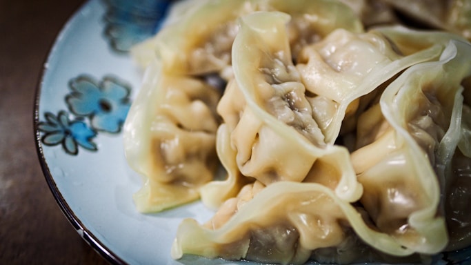A plate of dumplings is arranged on a decorative dish with floral patterns. The dumplings have a smooth, glossy texture and appear freshly cooked, with steam visible rising from them.
