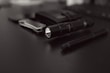 A collection of everyday carry items placed on a dark surface includes a flashlight, a folding knife, a pen, and a wallet. The focus is on the black flashlight with a textured grip, while the other items are slightly blurred in the background.