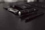 A collection of everyday carry items placed on a dark surface includes a flashlight, a folding knife, a pen, and a wallet. The focus is on the black flashlight with a textured grip, while the other items are slightly blurred in the background.
